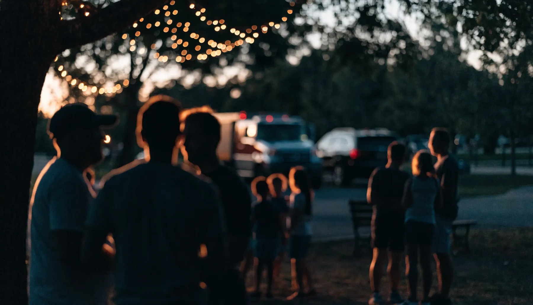 Families and first responders gathered as silhouettes at twilight under string lights in a park